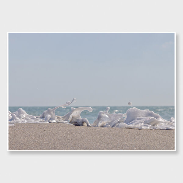 Foam Dance art print by Crystal May Photography. Close-up of ocean foam captured mid-motion on sandy beach under soft blue sky. Coastal fine art photograph with playful, sculptural wave forms and natural light.