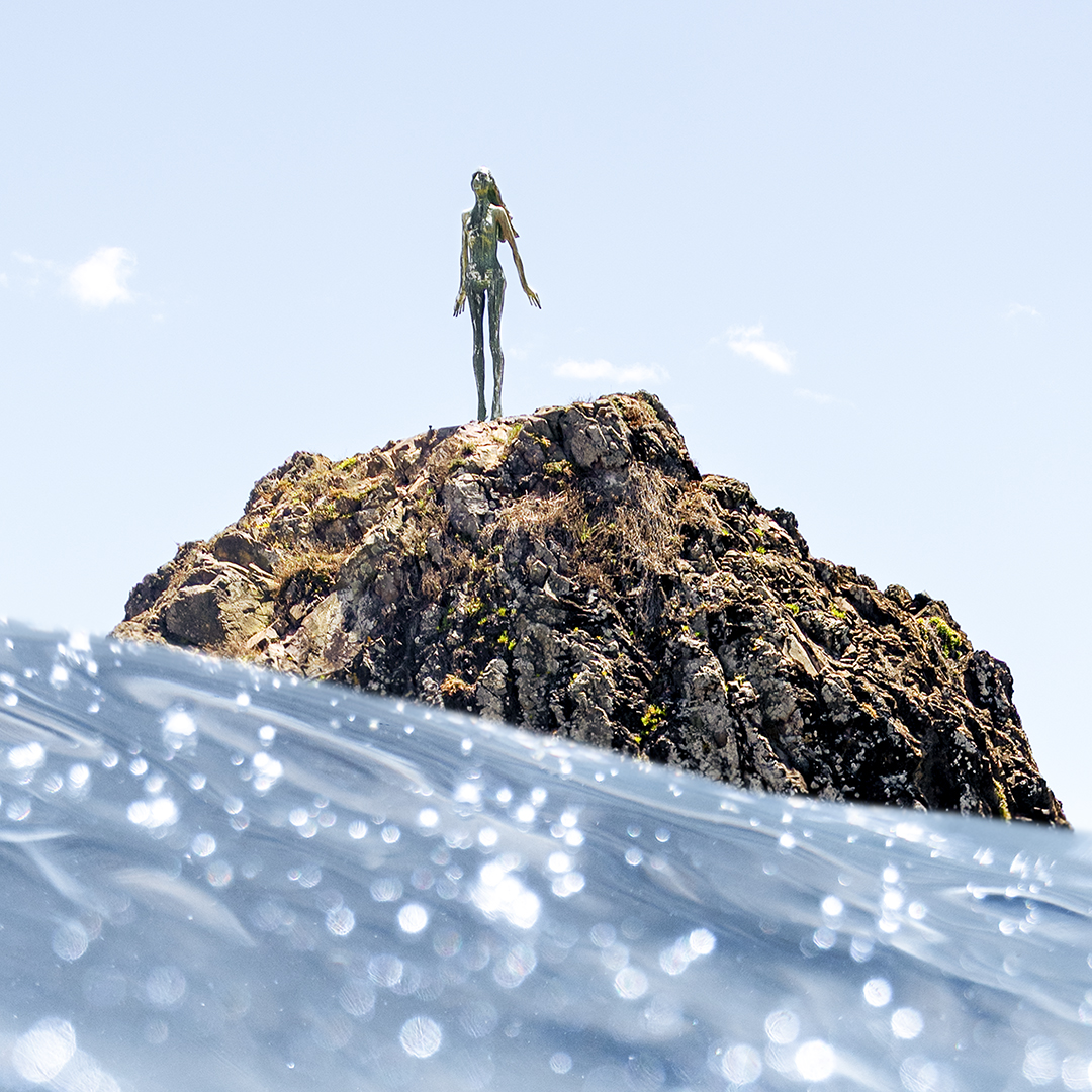 Fine art ocean photograph of The Lady on the Rock in Whakatāne, New Zealand, photographed from the water with sparkling blue ocean in the foreground and the sculpture standing above a rocky outcrop.